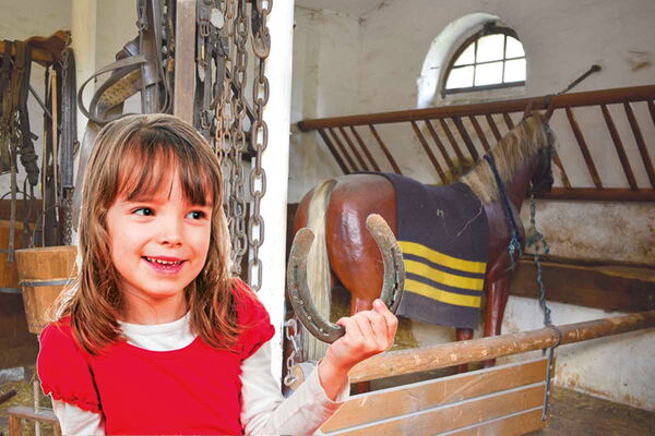 Ein Mädchen mit dunkelblonden Haaren und einem roten T-Shirt hält ein Hufeisen in der Hand. Hinter ihr ist die Ausstellung im Kommandantenpferdestall mit einem Holzpferd, einer Futterraufe und weiteren Ausrüstungsstücken.