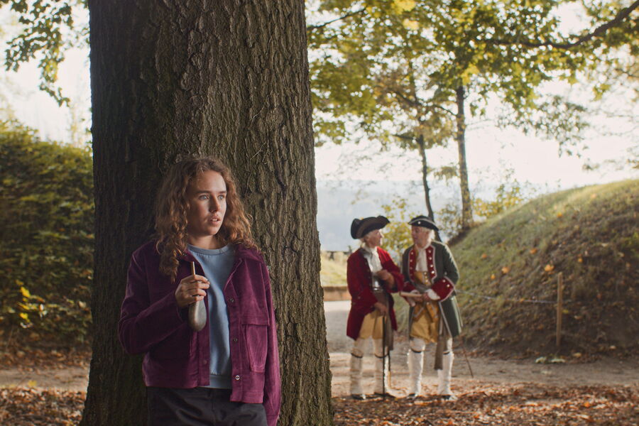 Standbild aus der Filmprojektion: ein junges Mädchen mit einer Pulverflasche in der Hand belauscht ein Gepräch zwischen historisch uniformierten Herren