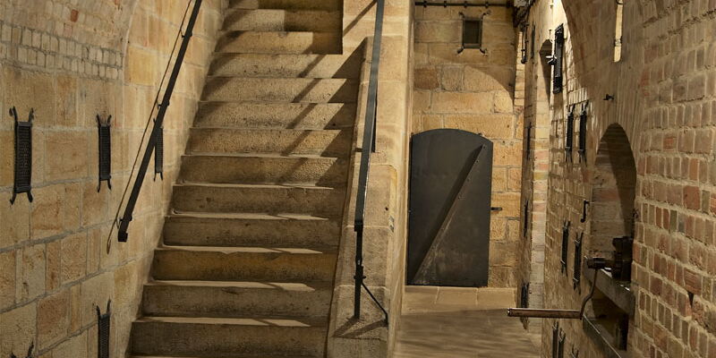 In einem Gewölbe aus Sandstein sind mehrere Klappen und Gitter sowie die Kurbel eines Munitionsaufzuges. Hinten links ist eine Treppe. Hinten rechts steht eine Metalltür offen.