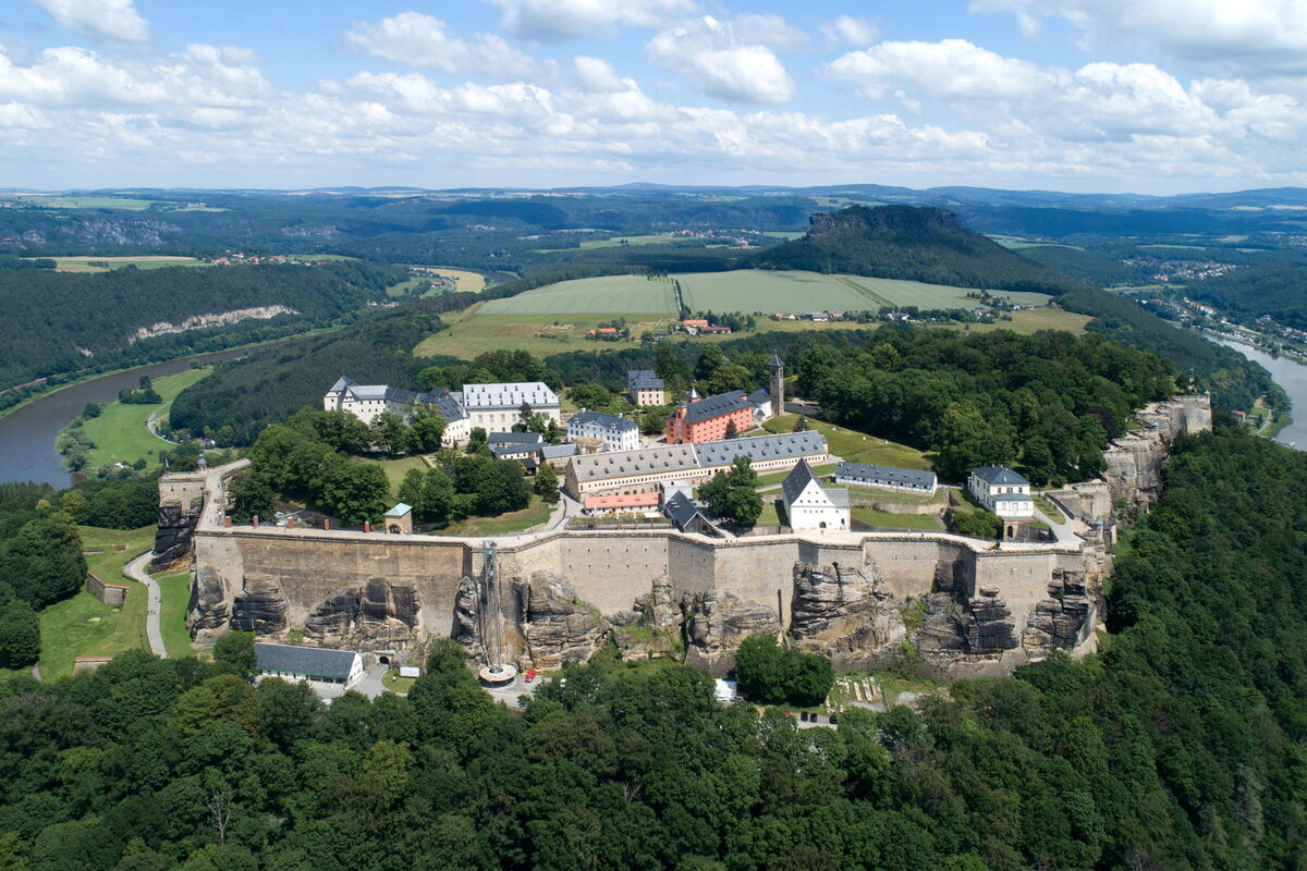 Eine Luftaufnahme zeigt die Festung Königstein. Im Hintergrund sind der Lilienstein, die Elbe und weitere Teile der Sächsischen Schweiz. Der Himmel ist blau und von Wolken durchzogen.