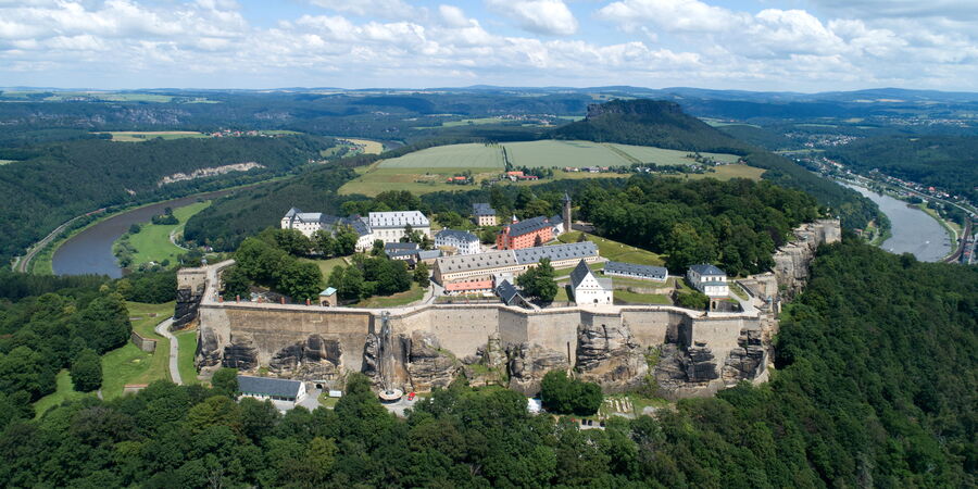 Letecký pohled ukazuje pevnost Königstein. V pozadí je vidět Lilienstein, Labe a další části Saského Švýcarska. Obloha je modrá a protkaná mraky.