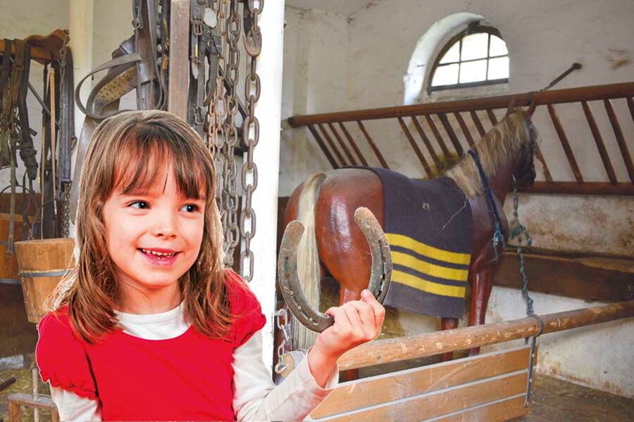 Ein Mädchen mit dunkelblonden Haaren und einem roten T-Shirt hält ein Hufeisen in der Hand. Hinter ihr ist die Ausstellung im Kommandantenpferdestall mit einem Holzpferd, einer Futterraufe und weiteren Ausrüstungsstücken.