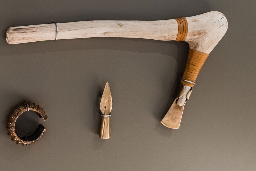 Auf einem dunklen Untergrund liegen drei Nachbildungen historischer Gegenstände.  Links liegt ein Armreif mit groben Verzierungen aus Metall. In der Mitte liegt eine Pfeilspitze. Rechts liegt eine Nachbildung einer Axt aus der Bronzezeit mit einem Holzgri